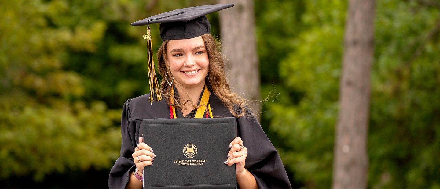 A female Oakland University graduate, wearing a cap and gown, holding up a diploma.